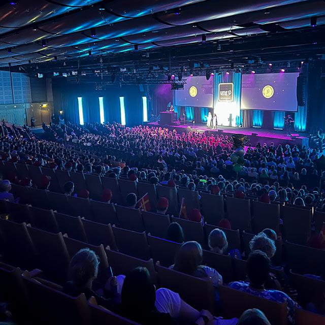 A packed auditorium with an audience watching a lit stage during an event.