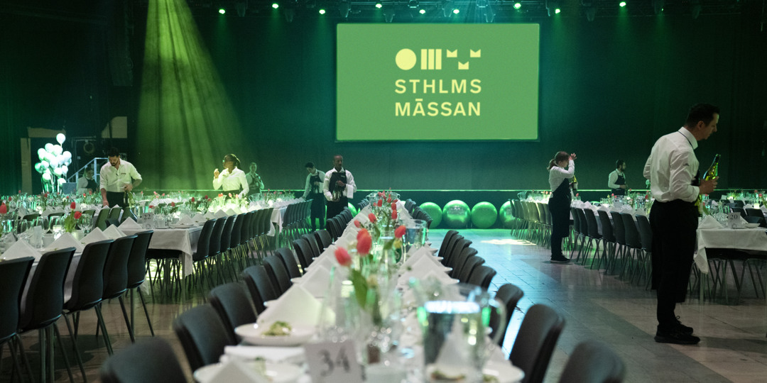 victoriahallen-stockholmsmässan-kostdagarna-2025 Sittande långbordsdukning i Victoriahallen. Röda tulpaner på borden och personal som håller på att duka inför middagen. En grön skärm visar Stockholmsmässans logotyp i gult.