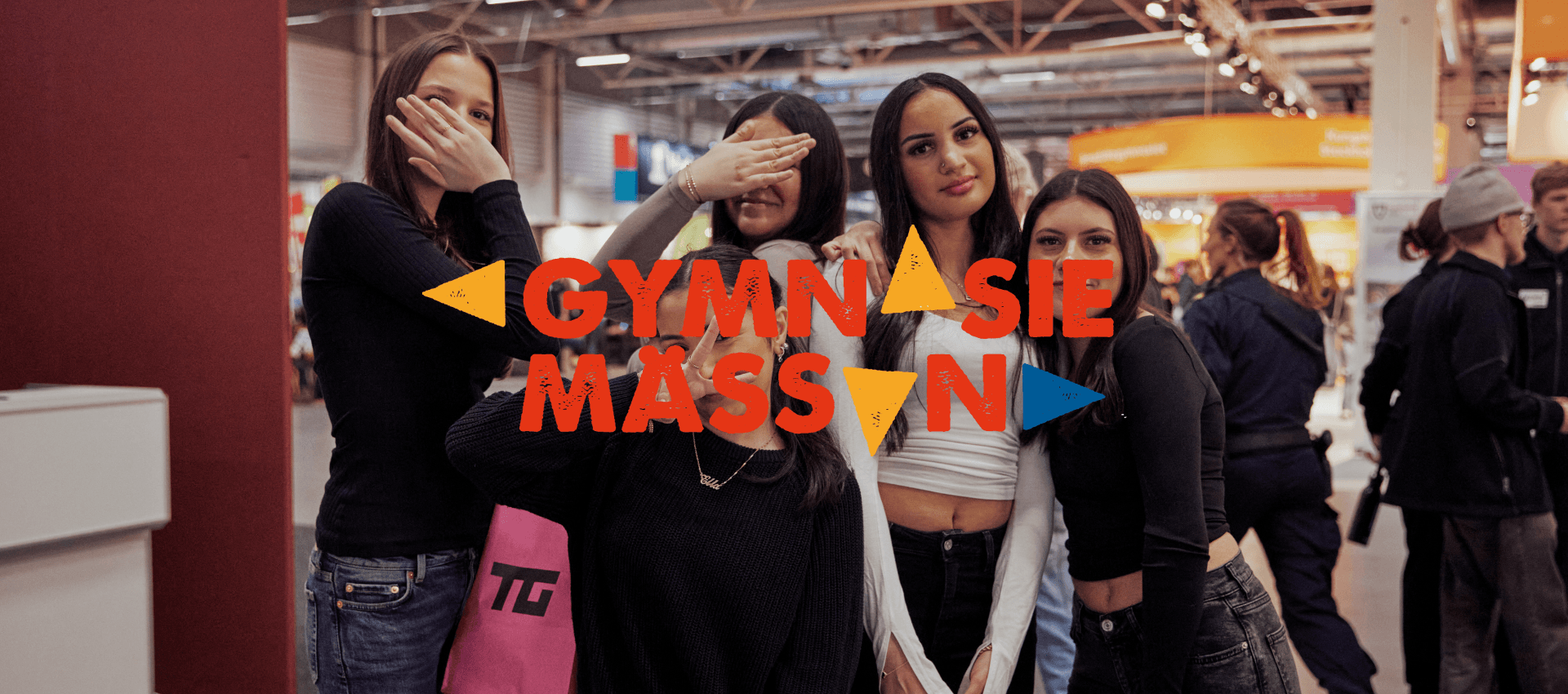 A group of five people are standing together at a fair. The text "GYMNASIEMÄSSAN" is placed over the image in red color with yellow and blue triangles around the text.