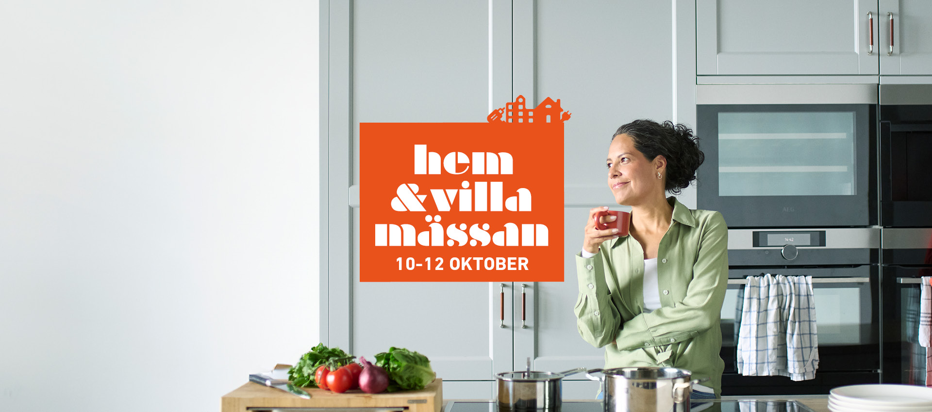 En kvinna står i ett modernt kök och dricker från en kopp. Framför henne finns en köksö med grönsaker och kastruller. En orange skylt med texten "hem & villa mässan 10-12 OKTOBER" är placerad över bilden.