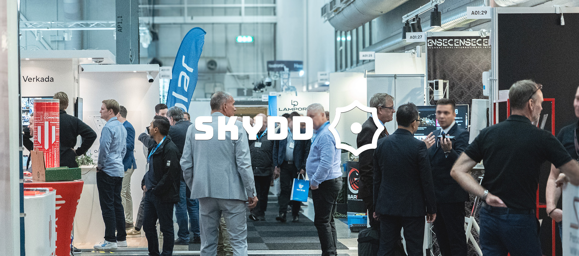 A trade fair with several booths and people walking around looking at exhibits. The text "SKYDD" is visible in the center of the image.