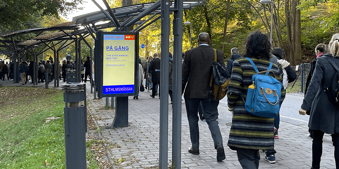 Inför ditt besök 2 Människor går längs en gångväg med skyddande tak mot Stockholmsmässan. Träd och en digital skylt syns i bakgrunden.