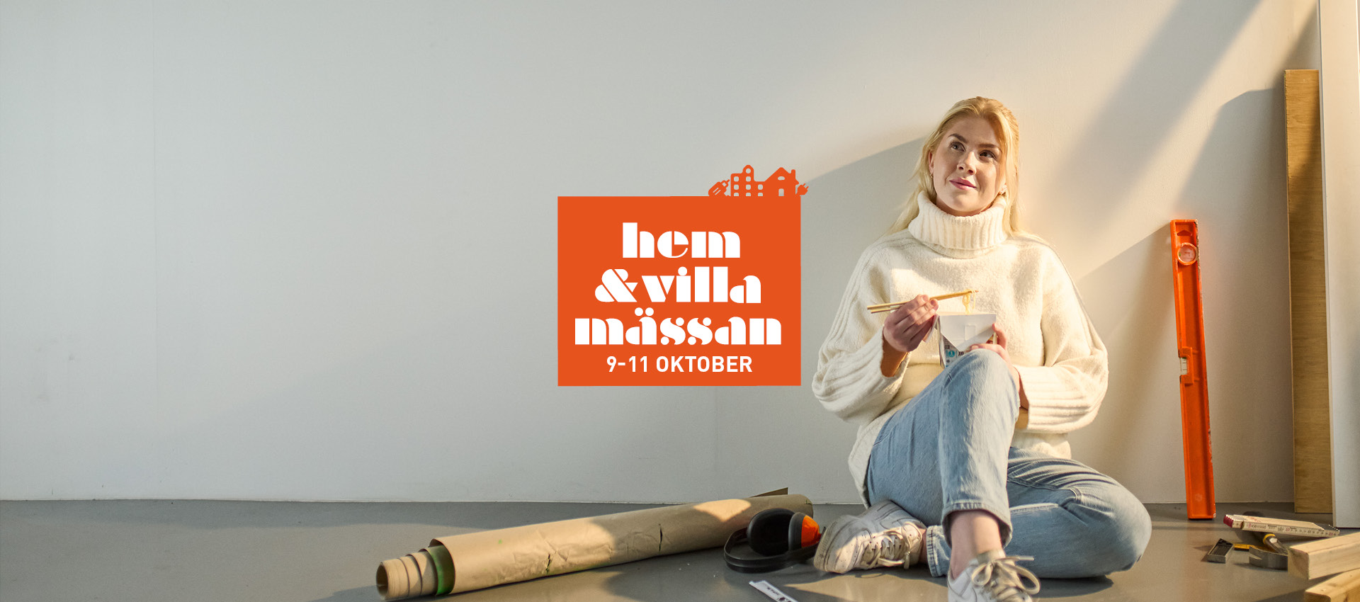 A person is sitting on the floor painting a wall. The person is wearing a white sweater and blue jeans. On the floor, there is a roll of paper, a spirit level, ear protection, and some wooden pieces. In the background, there is text that reads "hem & villa mässan 9–11 OKTOBER" in white against an orange background