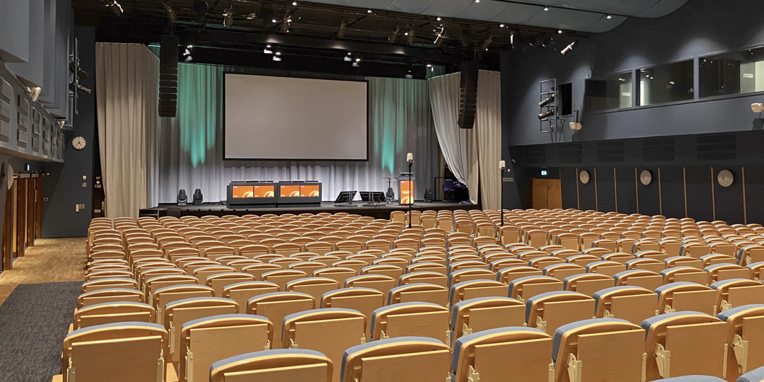 K1-lokaler-stockholmsmässan-stockholm-1080x540px.jpg Tom föreläsningssal med scen, projektorduk och stolsrader inför kommande presentation.