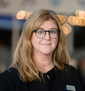Porträtt av en kvinna med axellångt, lockigt hår, svarta glasögon och svart blus.