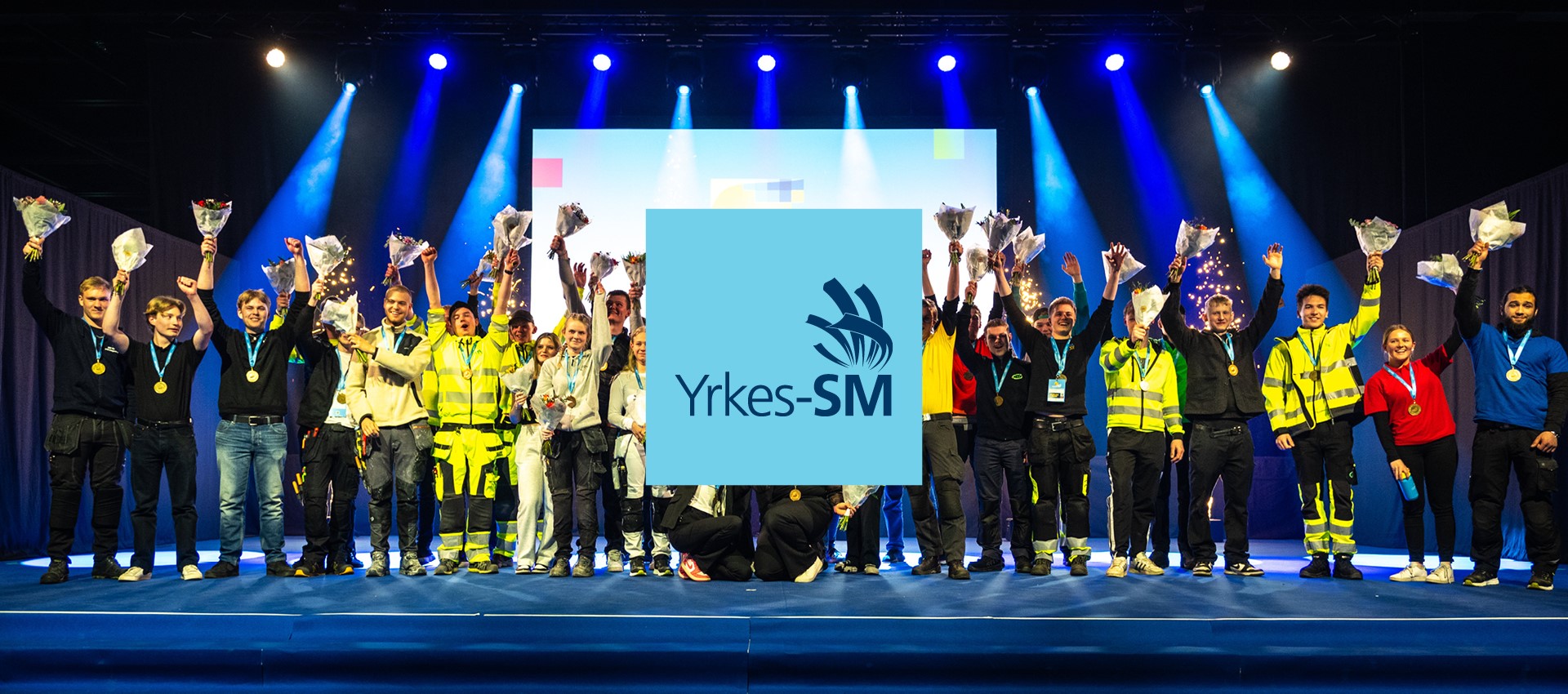 En grupp människor står på en scen med blå belysning och håller upp blommor och medaljer. De är klädda i olika arbetskläder och uniformer. I mitten av bilden finns en logotyp för Yrkes-SM.