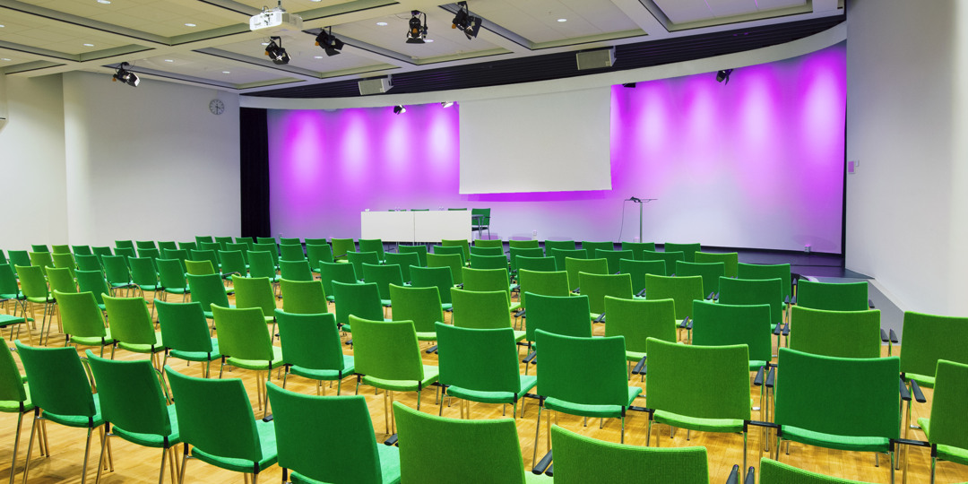 Färgstark konferenssal med gröna stolar och scen belyst i lila, redo för presentation.