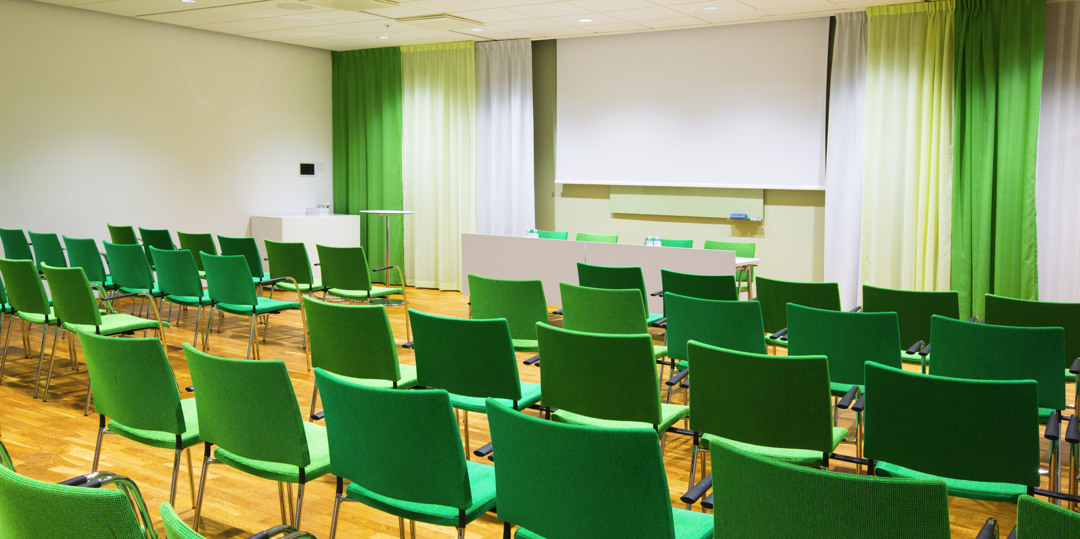 Ljus konferenslokal med gröna stolar och scen, förberedd för seminarium eller möte.