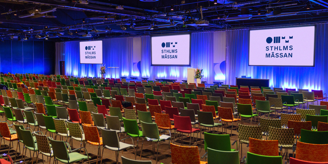T1-T2-3-screens-stockholmsmässan-konferensrum-1080x540px.jpg Stort konferensrum med färgstarka stolar och tre skärmar, förberett för evenemang på Stockholmsmässan.