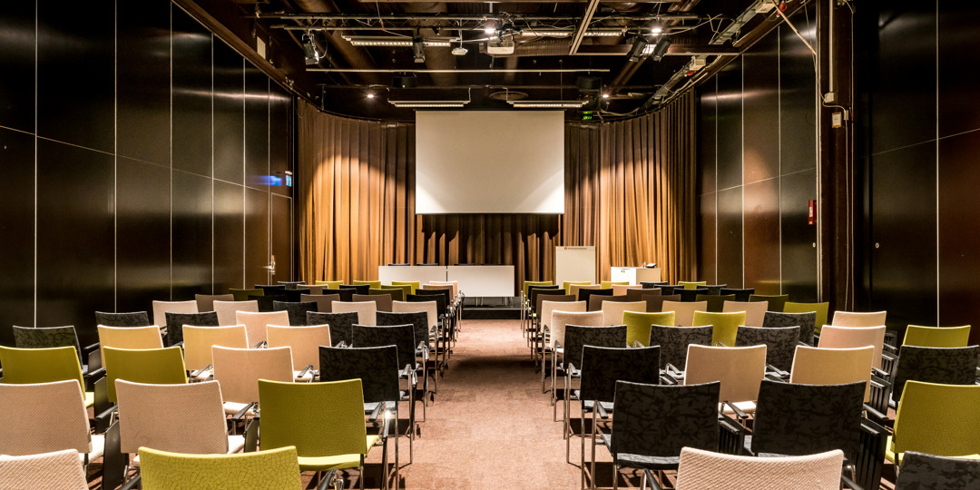 Konferensrum T4 med neutralt färgade stolar, scen och projektorduk, förberett för föreläsning.