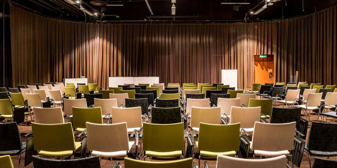 Konferensrum med scen och varierat färgsatta stolar, redo för föreläsning eller seminarium.