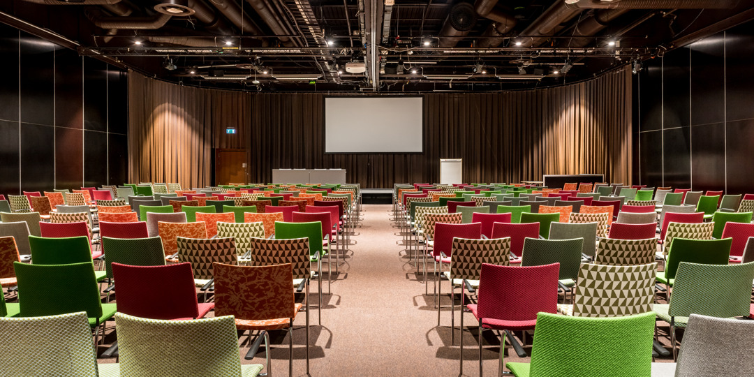 Konferenssal med färgglada stolar, scen och projektorduk, redo för föreläsning eller event.