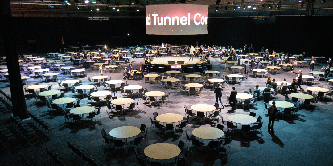 world-tunnel-congress-2025-Think-tank-stockholm-fair-1080x540px Large round tables surround a central stage, the Think Tank, in one of Stockholmsmässan's halls during an ongoing congress.