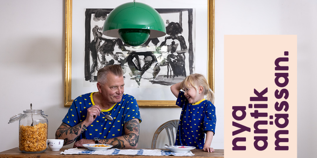 En person och ett barn sitter vid ett köksbord och äter frukost med flingor. På bordet står en stor glasburk med cornflakes och två skålar. De bär matchande blå tröjor med gula mönster. I bakgrunden syns en tavla med svartvit konst och en grön taklampa hänger ovanför bordet. Till höger i bilden finns en beige yta med texten ”nya antik mässan.” i mörklila bokstäver.