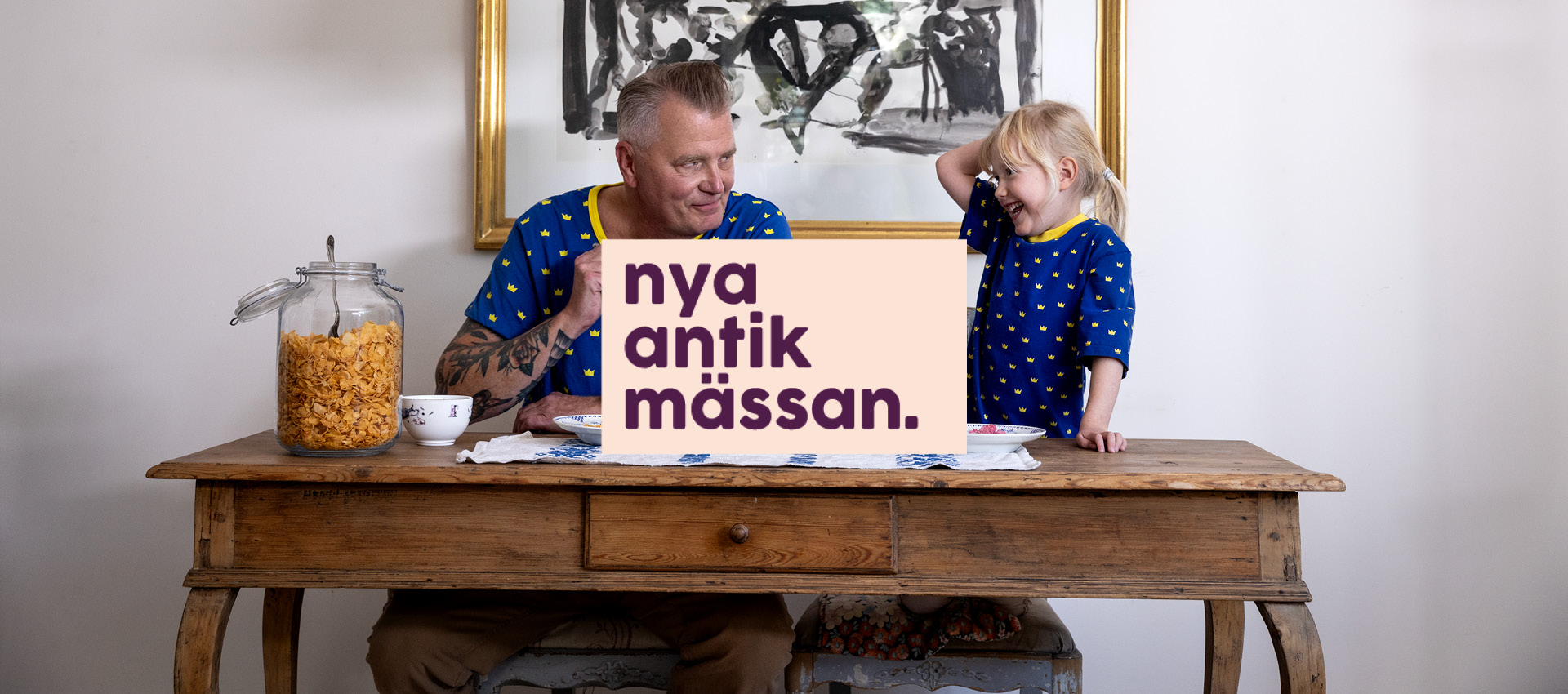 En person och ett barn sitter vid ett köksbord och äter frukost med flingor. På bordet står en stor glasburk med cornflakes och två skålar. De bär matchande blå tröjor med gula mönster. I bakgrunden syns en tavla med svartvit konst och en grön taklampa hänger ovanför bordet. Till höger i bilden finns en beige yta med texten ”nya antik mässan.” i mörklila bokstäver.