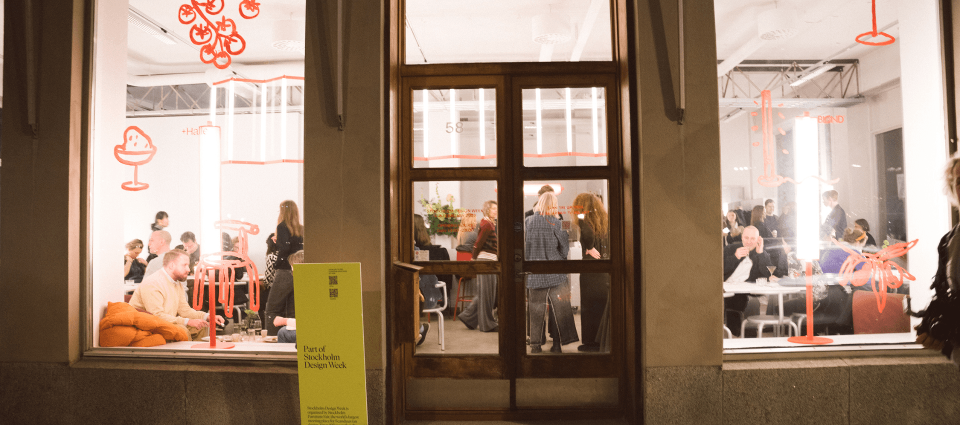 Ett kvällsevenemang sett utifrån genom stora fönster, där flera personer sitter och står runt bord inne i en ljus lokal. Väggarna är dekorerade med röda grafiska illustrationer, och en gul skylt vid dörren visar texten "Part of Stockholm Design Week".