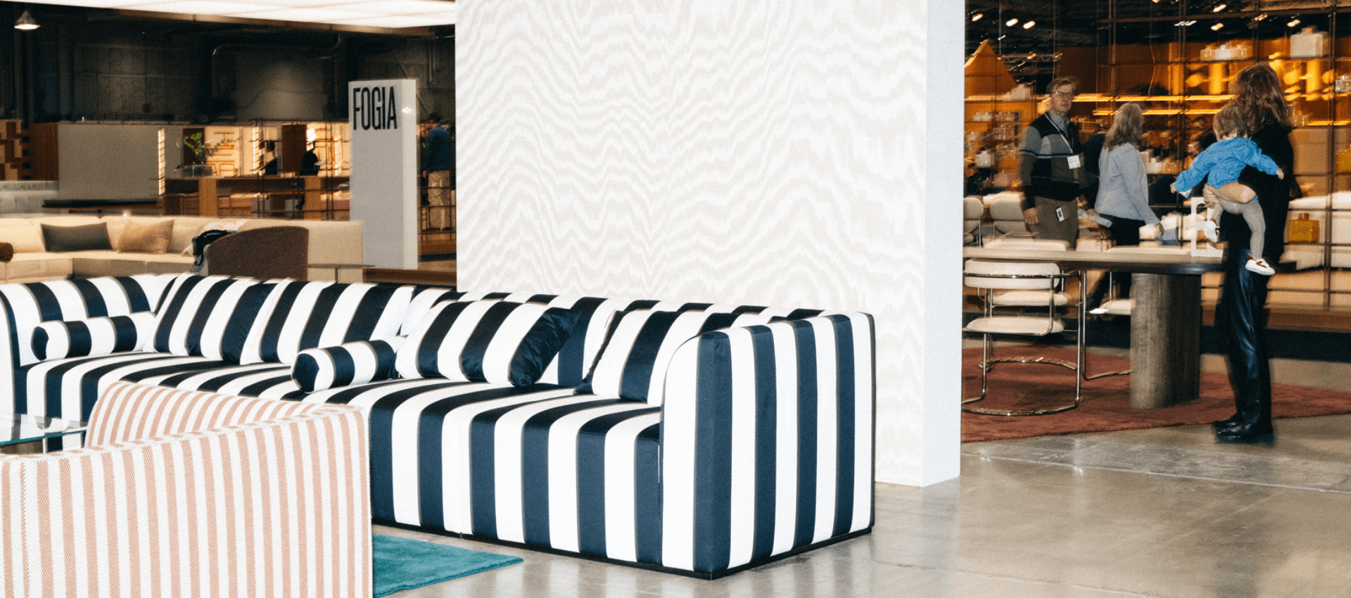 En modern utställningsmiljö med en stor soffa i svartvita ränder och en fåtölj med ljusröda och vita ränder i förgrunden. I bakgrunden syns ett bord med stolar och människor som samtalar, samt en skylt med texten "FOGIA". Golvet är av betong och belysningen är industriell.