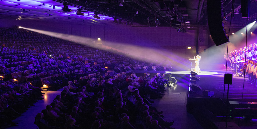 Full audience watching a concert with singer and choir in vibrant lighting inside Victoria Hall at Stockholmsmässan.