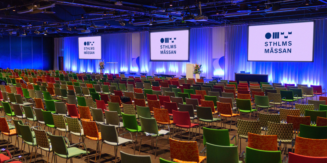 Colourful conference room with stage, three large screens and the Stockholmsmässan logo.