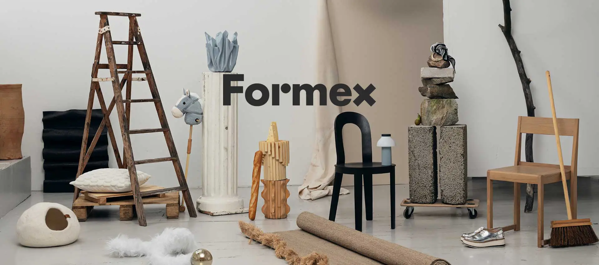 En stylad inomhusscen med olika inrednings- och designobjekt, där Formex‑logotypen syns i mitten; bland objekten finns en trästege, stolar, pedestaler, dekorativa föremål och naturmaterial arrangerade i ett kreativt stilleben.