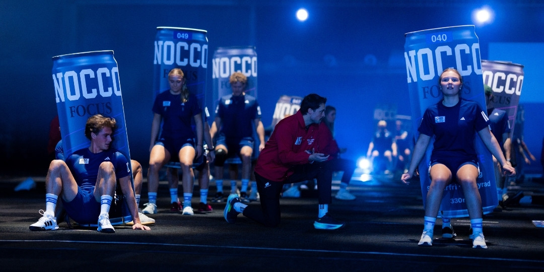 Deltagare i NOCCO Games utför ett tävlingsmoment sittande framför stora NOCCO-burkar i en upplyst eventmiljö.