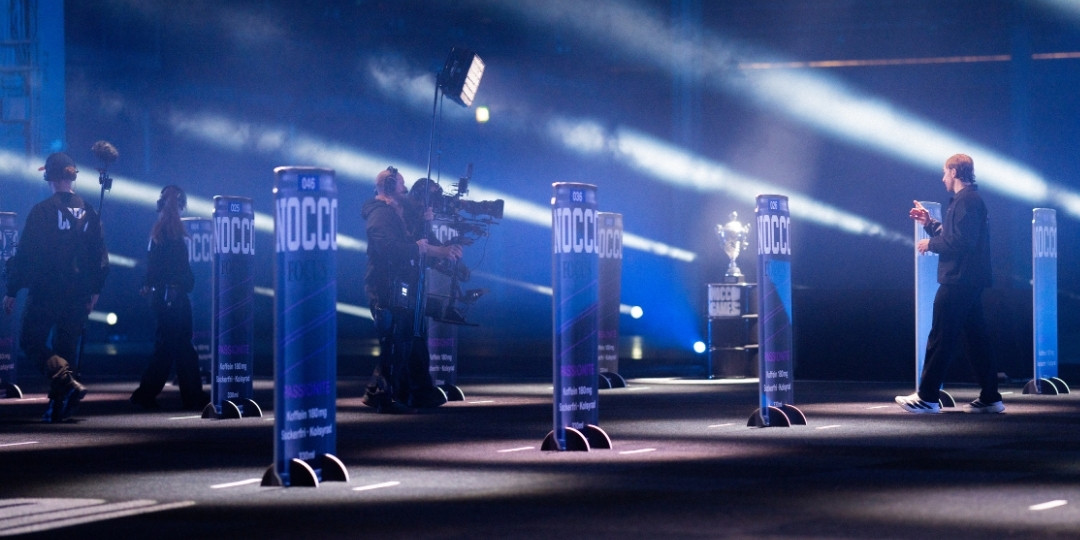 Kameraproduktion under NOCCO Games med filmteam, deltagare och programledare i en ljussatt eventmiljö med NOCCO-pelare.
