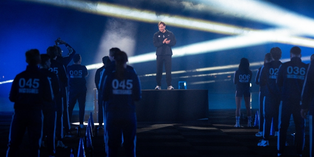 Deltagare i NOCCO Games står uppställda runt en scen där en person står i centrum, med ljuseffekter i en mörk eventmiljö.