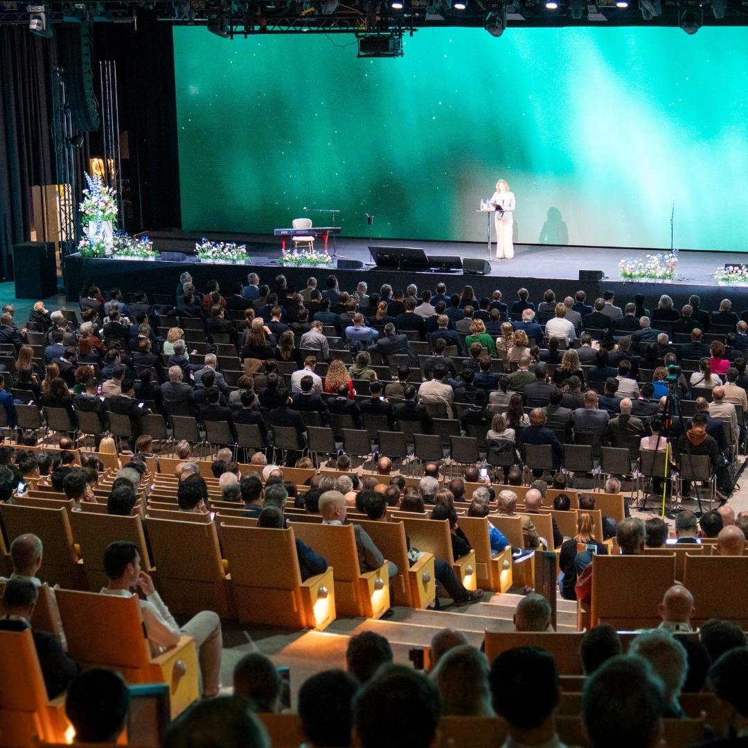 En rymlig evenemangslokal med färgglad belysning och en stor LED-skärm som visar en bild på fåglar. Två personer går över det öppna golvet, och till vänster syns sittplatser i nivåer. A spacious event hall with colourful lighting and a large LED screen showing an image of birds. Two people are walking across the open floor, and tiered seating is visible on the left.