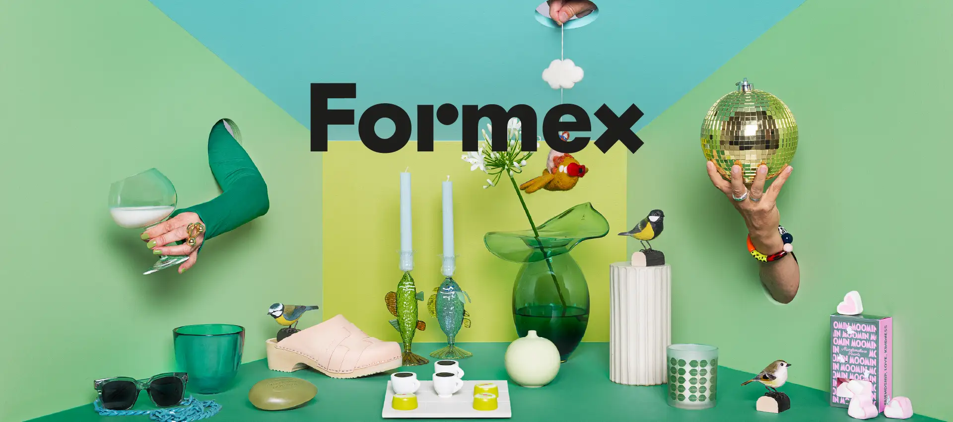 Färgstarkt stilleben för Formex med gröna inredningsdetaljer, dekorativa fåglar, blommor, ljusstakar, en spegelkula samt händer som sträcker sig genom väggen.